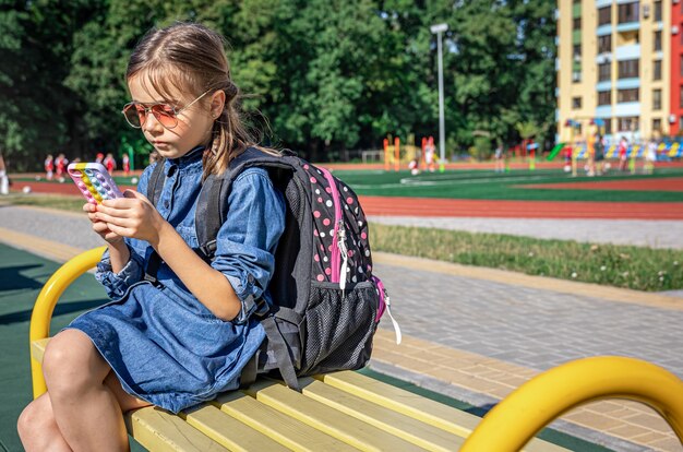 Un élève du primaire avec un sac à dos, utilise un smartphone, assis près de l'école.