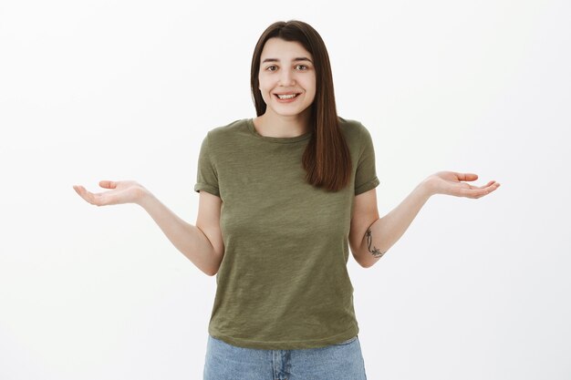 Elle n'en a aucune idée. Portrait of cute and friendly attractive young 20s caucasian girl making shoudler shrug comme écarté les mains sur le côté dans la pose désemparée, souriant stupide étant uanware sur mur gris