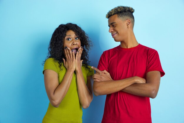 Elle a peur, il rit. Jeune bel homme afro-américain émotionnel et femme dans des vêtements colorés sur un mur bleu.