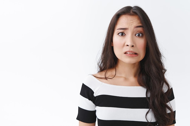 Embarrassée et confuse, une jeune femme asiatique peu sûre d'elle a fait une erreur se sentir désolée ou avoir pitié grincer des dents nerveusement et tirer un visage triste et inquiet froncer les sourcils paniquer debout fond blanc