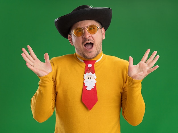 Photo gratuite Émotionnel jeune homme à col roulé jaune et lunettes avec cravate rouge drôle criant avec les bras levés debout sur le mur vert