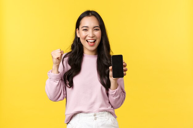 Les émotions des gens, les loisirs de style de vie et le concept de beauté. Fille asiatique mignonne excitée et heureuse chantant, pompe à poing triomphant et montrant l'affichage du téléphone portable, recommander une application ou un jeu pour smartphone.