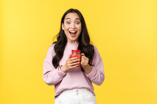 Les émotions des gens, les loisirs de style de vie et le concept de beauté. Une jolie fille asiatique excitée et heureuse entend des nouvelles géniales, tenant une boisson au smoothie et souriant émerveillée par la caméra, debout sur fond jaune