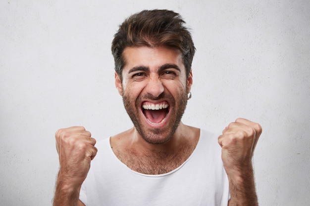 Émotions Positives, Victoire, Triomphe, Concept De Bonheur. Heureux Homme Avec Barbe Vêtu D'un T-shirt Blanc Décontracté, Serrant Les Poings Tout En Se Réjouissant De Sa Victoire De Jeu Posant Sur Blanc