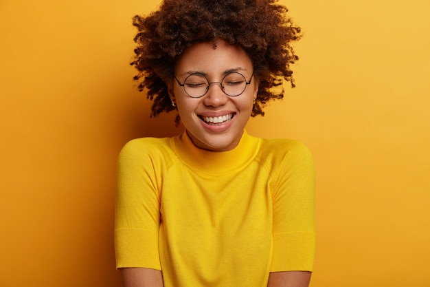 Photo gratuite Émotions pures et bonheur sincère. adolescente heureuse sourit, glousse à quelque chose de drôle, garde les yeux fermés, a le sourire à pleines dents, impressionnée par de merveilleuses nouvelles géniales, porte des vêtements jaunes