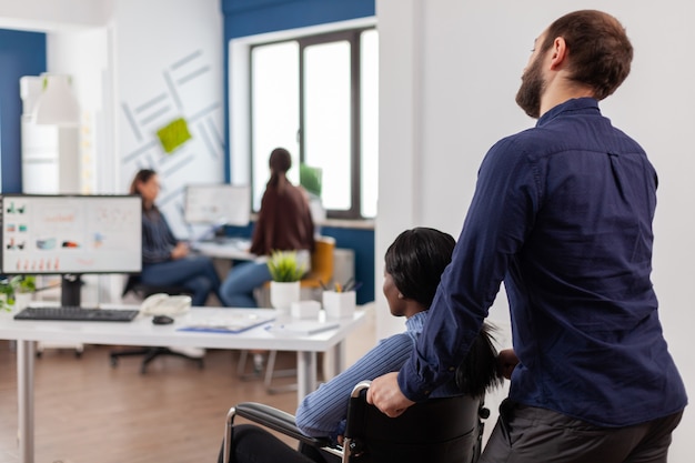 Un employé aide son collègue africain invalide à venir sur le lieu de travail en poussant un fauteuil roulant près du bureau