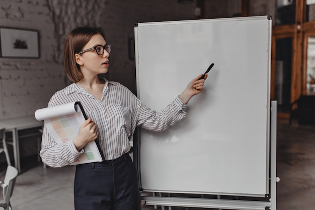 Photo gratuite employé aux cheveux courts en chemisier blanc et pantalon noir montre à bord du bureau. portrait de femme avec des documents sur les plans.