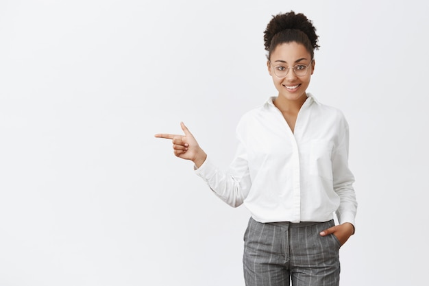 Employé de bureau charmant et sympathique montrant le chemin au client. Portrait de femme employeur intelligente et créative polie dans des lunettes et des pantalons, tenant la main dans la poche, pointant vers la gauche, indiquant à la sortie