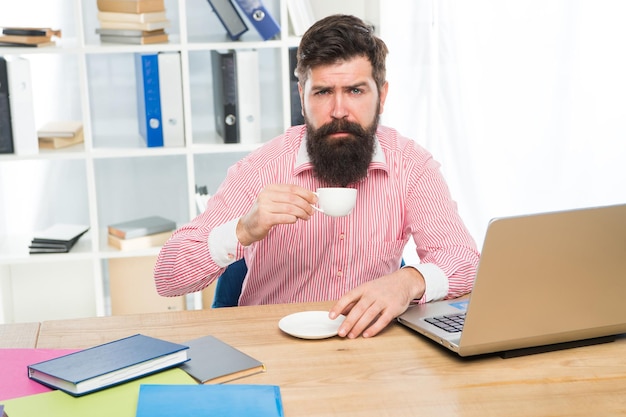 Employé de bureau épuisé avec barbe hipster boire du café assis au bureau dans un bureau moderne, le matin.