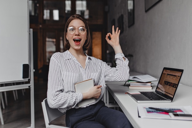 Employé de bureau fille joyeuse montre signe OK. Portrait de femme en pantalon et chemisier léger sur le lieu de travail.