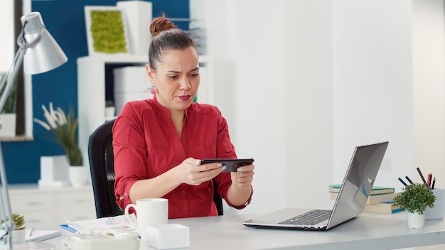 Employé de bureau jouant à des jeux vidéo en ligne sur smartphone, assis au bureau avec un ordinateur portable. Employé de l'entreprise s'amusant avec la compétition de jeux dans le cyberespace sur téléphone mobile dans un lieu de travail de démarrage.