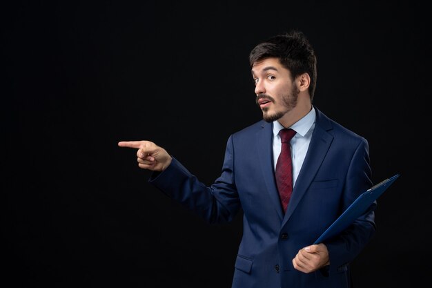 Employé de bureau masculin confus en costume tenant des documents et pointant quelque chose sur le côté droit sur un mur sombre isolé