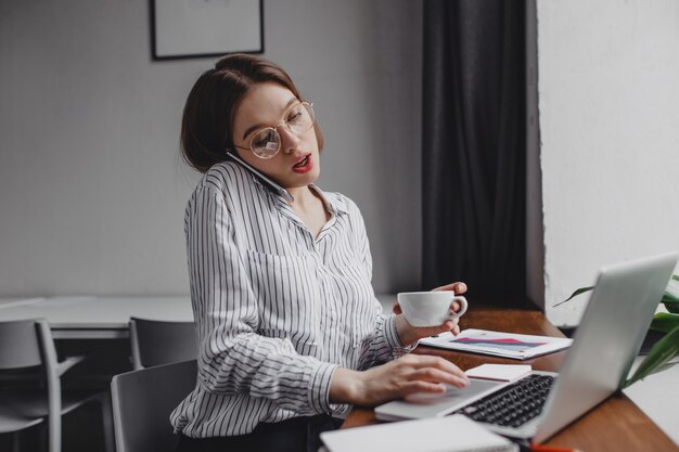 Employé de bureau occupé à parler au téléphone et à travailler dans un ordinateur portable, tenant une tasse de thé.