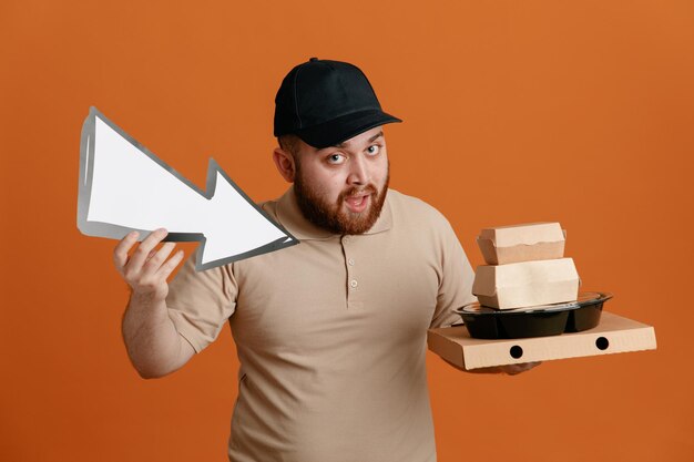 Employé de livreur en casquette noire et uniforme de t-shirt blanc tenant une flèche et des récipients alimentaires regardant la caméra debout heureux et positif sur fond orange