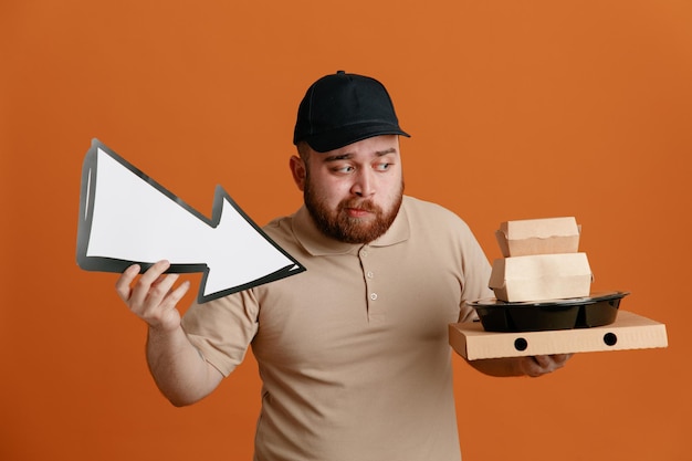 Photo gratuite employé de livreur en casquette noire et uniforme de t-shirt blanc tenant une flèche et des récipients alimentaires regardant de côté confus ayant des doutes debout sur fond orange