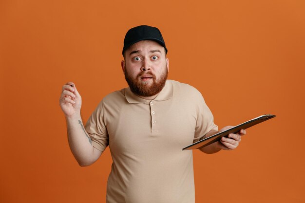 Employé de livreur en casquette noire et uniforme de t-shirt blanc tenant un presse-papiers avec un stylo regardant la caméra confus et surpris debout sur fond orange