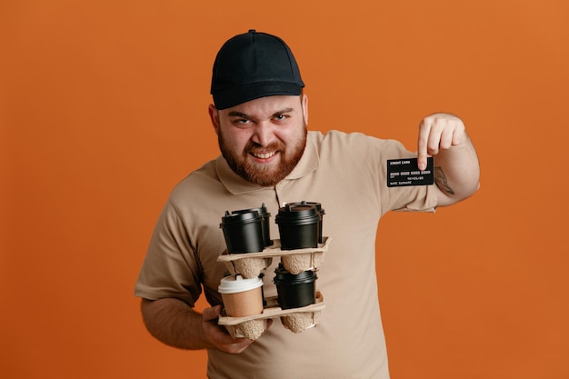 Employé de livreur en casquette noire et uniforme de t-shirt blanc tenant des tasses à café montrant une carte de crédit en regardant la caméra heureux et positif souriant joyeusement debout sur fond orange