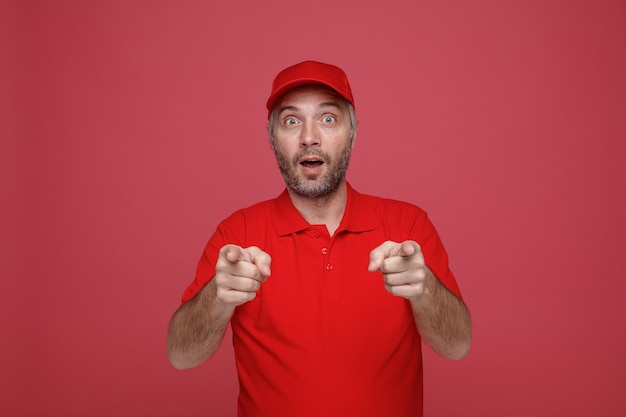 Employé de livreur en uniforme de t-shirt blanc à capuchon rouge pointant avec l'index vers la caméra heureux et surpris debout sur fond rouge