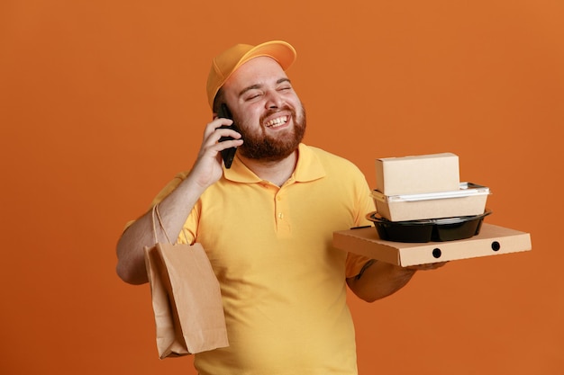 Employé de livreur en uniforme de t-shirt blanc à casquette jaune tenant un récipient de nourriture et une boîte à pizza avec un sac en papier parlant sur un téléphone portable souriant largement debout sur fond orange