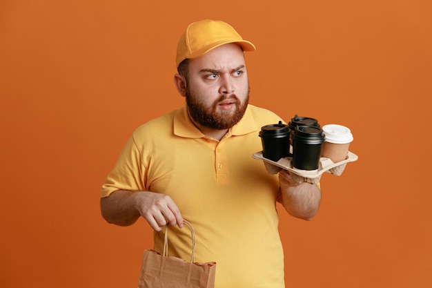 Employé de livreur en uniforme de t-shirt blanc à casquette jaune tenant des tasses à café et un sac en papier regardant de côté avec le visage fronçant les sourcils debout sur fond orange