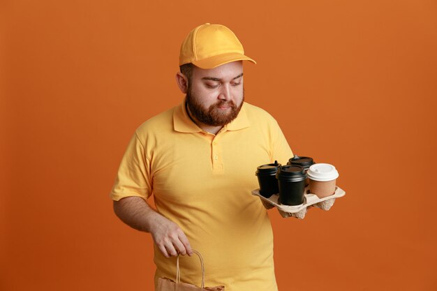 Employé de livreur en uniforme de t-shirt blanc à casquette jaune tenant des tasses à café et un sac en papier regardant des tasses avec un visage sérieux debout sur fond orange