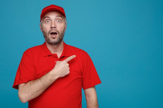 Employé de livreur en uniforme de t-shirt blanc à casquette rouge regardant la caméra étonné et surpris pointant avec l'index sur le côté debout sur fond bleu