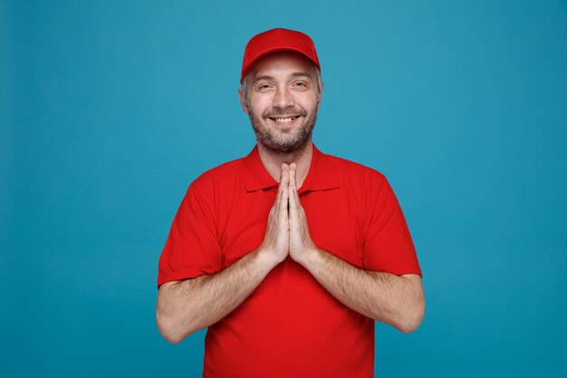 Employé de livreur en uniforme de t-shirt blanc à casquette rouge regardant la caméra heureux et heureux tenant les paumes ensemble souriant joyeusement debout sur fond bleu