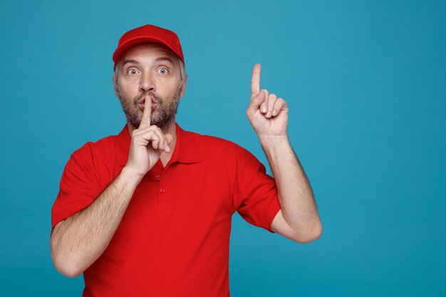 Employé de livreur en uniforme de t-shirt blanc à casquette rouge regardant la caméra surpris en faisant un geste de silence avec le doigt sur les lèvres pointant avec l'index vers le haut debout sur fond bleu