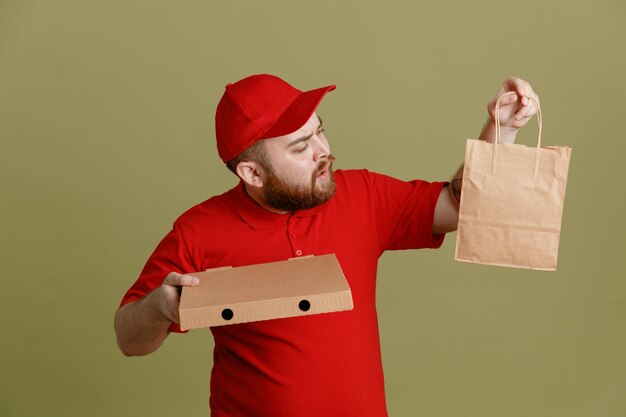 Employé de livreur en uniforme de t-shirt blanc à casquette rouge tenant une boîte à pizza et un sac en papier en le regardant être confus debout sur fond vert