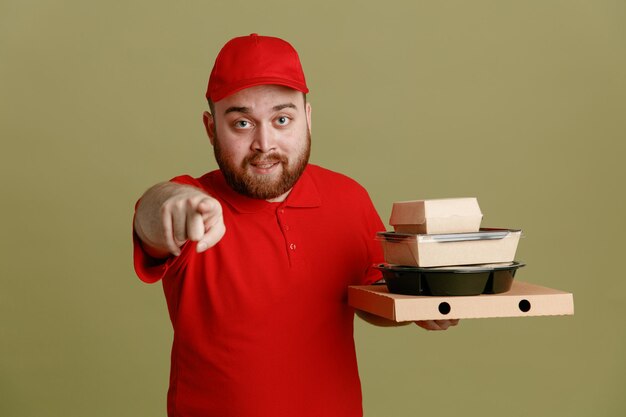 Employé de livreur en uniforme de t-shirt blanc à casquette rouge tenant des contenants de nourriture et une boîte à pizza pointant avec l'index vers la caméra souriant amical debout sur fond vert