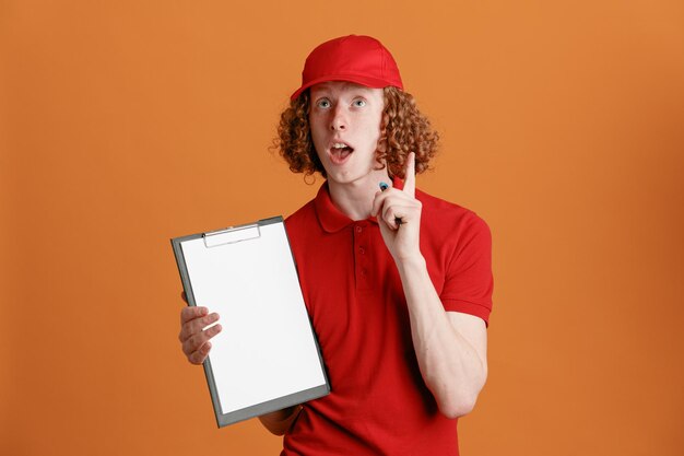 Photo gratuite employé de livreur en uniforme de t-shirt blanc à casquette rouge tenant un presse-papiers avec un stylo en attente de signature l'air surpris montrant l'index ayant une nouvelle idée debout sur fond orange