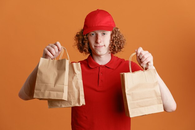 Employé de livreur en uniforme de t-shirt blanc à casquette rouge tenant des sacs en papier regardant la caméra heureux et positif souriant confiant debout sur fond orange