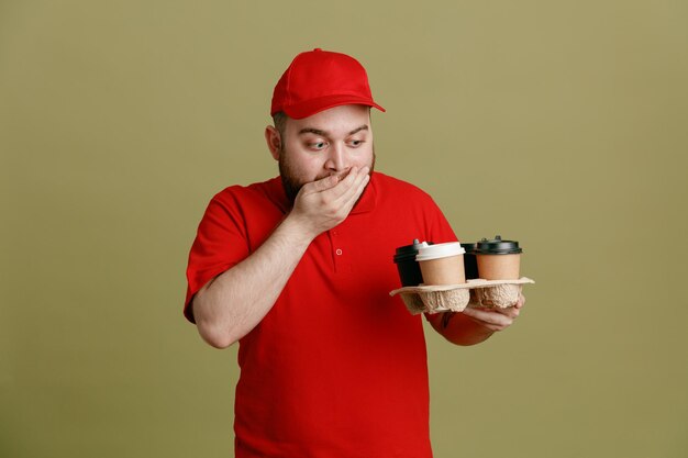 Employé de livreur en uniforme de t-shirt blanc à casquette rouge tenant des tasses à café en les regardant étonné et surpris couvrant la bouche avec la main debout sur fond vert