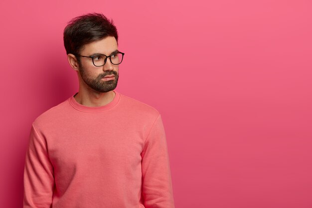 Un employé masculin barbu à l'air sérieux concentré sur le côté droit, a des pensées profondes sur les futurs problèmes de travail, porte des lunettes transparentes et un pull rose, pose à l'intérieur. Prise de vue monochrome