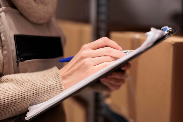 Photo gratuite employé de stockage du bureau de poste tenant le presse-papiers dans l'entrepôt, marquant la liste de contrôle de l'inventaire. employé d'entrepôt écrivant à la main avec un stylo en gros plan, vérifiant les boîtes en carton dans l'entrepôt