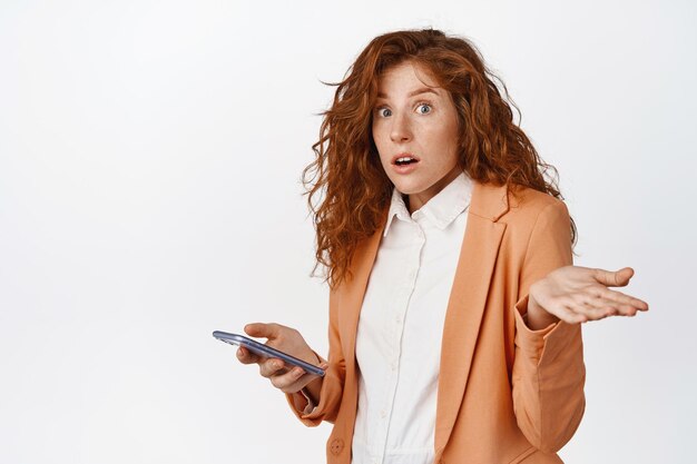 Employée de bureau rousse confuse tenant un téléphone portable en haussant les épaules et regardant la caméra désemparée debout en costume sur fond blanc