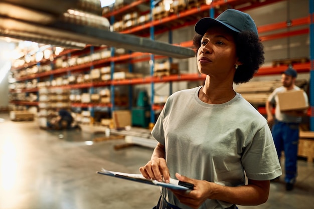 Employée d'entrepôt noire inspectant des marchandises dans un compartiment de stockage industriel