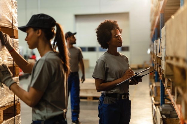 Employée d'entrepôt noire passant par la liste d'expédition tout en vérifiant le stock dans le compartiment de stockage industriel