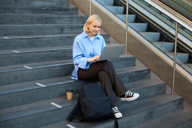 Photo gratuite employée indépendante travaillant sur son projet à l'aide d'un ordinateur portable assise à l'extérieur sur les escaliers à boire