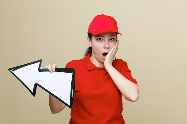 Photo gratuite employée de livraison en bonnet rouge et uniforme de t-shirt blanc tenant des récipients alimentaires et une grande flèche regardant la caméra étonnée et surprise debout sur fond marron