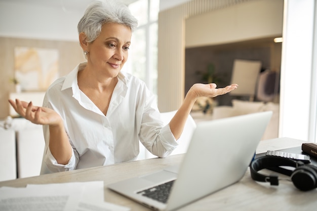 Employée mature émotionnelle en chemise blanche travaillant à domicile, assise à table avec un ordinateur portable, faisant un geste impuissant, haussant les épaules, ayant un chat virtuel en ligne
