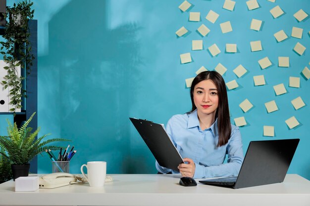 Employée d'une start-up vérifiant les e-mails sur un ordinateur portable et prenant des notes sur les tâches en attente. Femme assise au bureau moderne avec téléphone fixe et fournitures de travail sur la table.