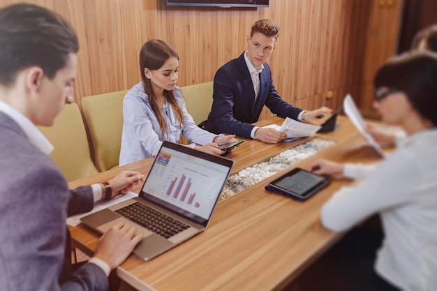 Les Employés De Bureau Tiennent Une Réunion à Un Bureau Pour Les Ordinateurs Portables, Les Tablettes Et Les Papiers, Un Grand Téléviseur Sur Un Mur En Bois