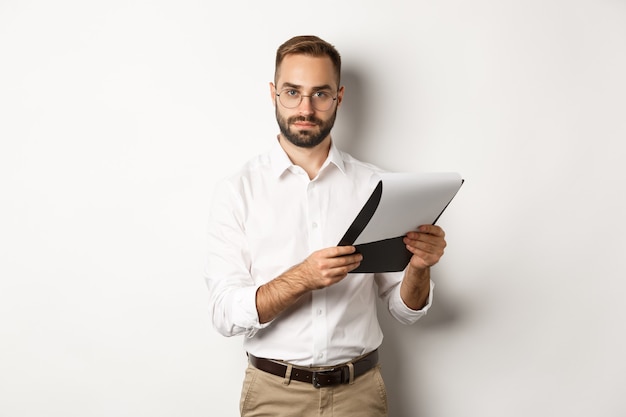 Employeur sérieux regardant la caméra tout en lisant des documents sur le presse-papiers, ayant un entretien d'embauche, debout