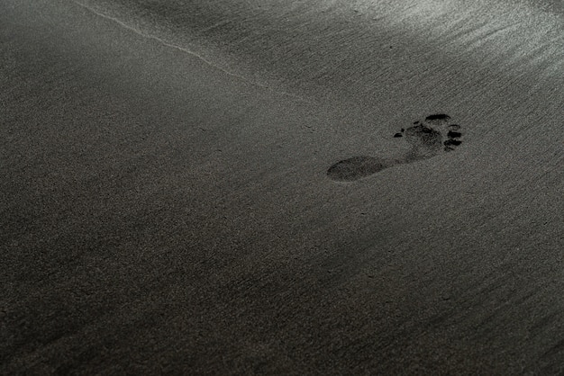 Empreinte sur une macro photographie de plage de sable noir. Trace humaine sur une texture de plage noire et soyeuse avec une faible profondeur de champ. Fond noir minimaliste. Ténérife rivage sablonneux voulcanic.