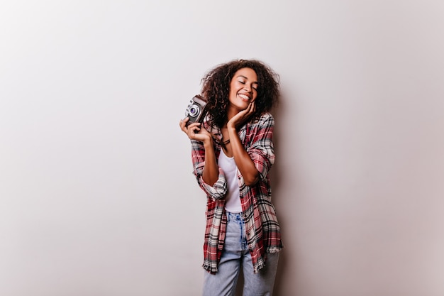 Enchanteur féminin heureux posant. Jolie dame africaine avec caméra debout sur blanc et riant.