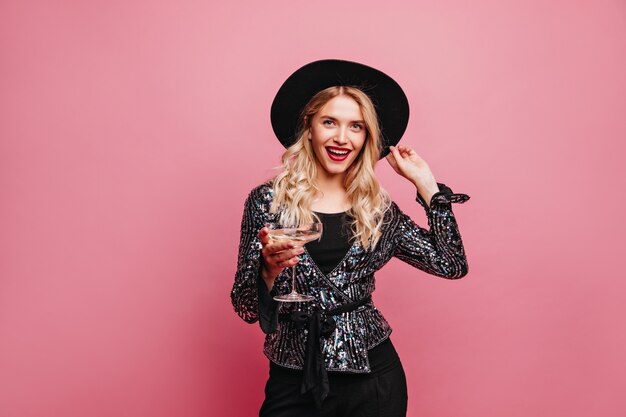 Enchanteur femme blanche posant avec verre à vin sur un mur pastel. Photo intérieure d'une fille blonde de bonne humeur au chapeau noir.