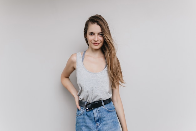 Enchanteur fille aux cheveux longs posant avec une expression de visage heureux. Portrait intérieur d'une jeune femme brune confiante en jupe en jean.