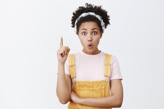 Photo gratuite encore une idée. excité séduisante femme à la peau sombre en bandeau et salopette jaune, levant l'index en geste eureka, pliant les lèvres et regardant tout en expliquant le grand plan qu'elle a inventé