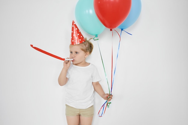 Enfance, bonheur, célébration et concept amusant. Adorable petit enfant soufflant sifflet, tenant des ballons colorés, se sentir heureux, célébrer l'anniversaire, posant au mur blanc